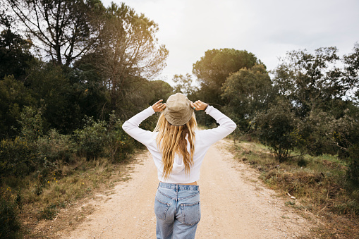 Rear view of a young beautiful blonde woman wearing a hat happily walking on a path in a forest