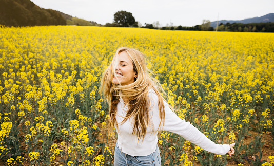Beautiful young blonde cheerful woman, wearing a hat, poses amidst a field of blooming yellow rapeseed flowers