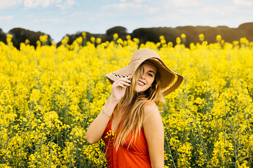 Portrait of a young beautiful blonde woman, wearing a red dress and a hat, poses amidst a field of blooming yellow rapeseed flowers
