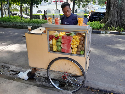June 3, 2023 - Bandung, West Java, Indonesia.
A man selling freshly cut tropical fruit from a traditional Indonesian food cart or grobak on the roadside in Bandung, West Java, Indonesia. The fruits include watermelon, pineapple, mango, melon, papaya, strawberries, apples and kiwi fruit.
