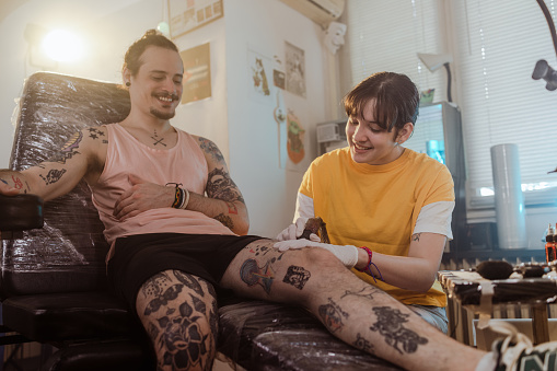 Portrait of a young woman with protective gloves tattooing a Latin American man in a tattoo parlor