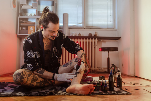 Portrait of a young Latin American man with protective gloves sitting on the floor and  tattooing his ankle