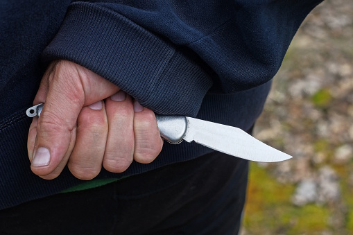 hand of a male criminal in blue clothes and a gray knife behind his back on the street