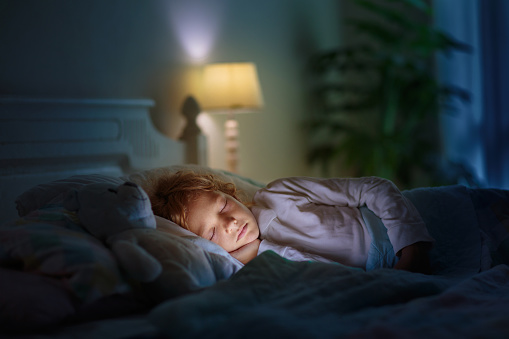 Child sleeping in dark bedroom. Little boy napping. Healthy night rest. Kids room with night light. Toddler with teddy bear. Bedding for children.