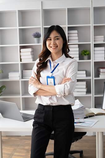 Portrait of young smiling Asian businesswoman looking at camera with crossed arms, standing in the office room.