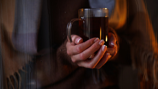 Close-up of glass cup with tea in female hands outside window wet from rain,drops flow down glass.Atmospheric photo autumn mood sadness,melancholy, loneliness.Autumn,fall,home comfort in cold season