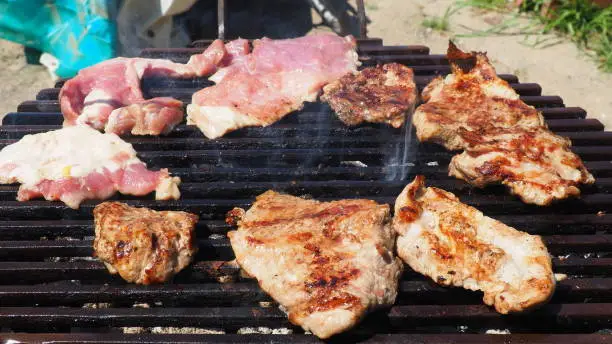 Grilled juicy meat pork steaks in burning coals on a barbecue grill, white smoke. Crispy crust. Tasty steak with blood and fried strips. Men's food. Grilled juicy striped steak is ready for a picnic.