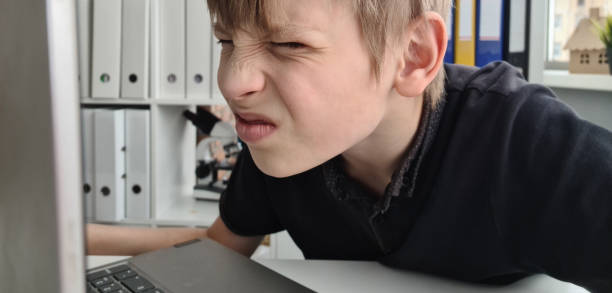 Teenager boy squints using a laptop with poor eyesight sits and looks at computer Teenager boy squints using a laptop with poor eyesight sits and looks at computer. Online student at home squint eyes stock pictures, royalty-free photos & images