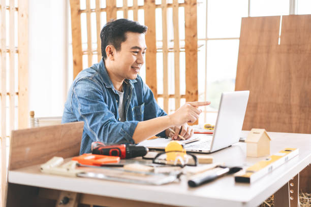 mittelalter erwachsener asiatischer mann tischler mit laptop für online-arbeit mit kunden wotk zu hause - southeast asian ethnicity men laptop image type stock-fotos und bilder