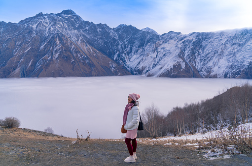 Happy woman tourist with fantastic scenery of the Caucasus mountains and sea of mist in winter near Gergeti Trinity Church in Kazbegi, Georgia.