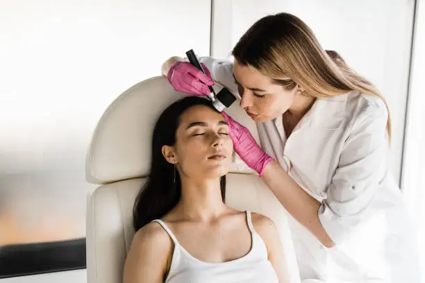 Photo of Dermatoscopy of skin lesions and moles with dermatoscope for preventing skin cancer and melanoma. Dermatologist examines moles and birthmarks of girl patient using dermatoscope.