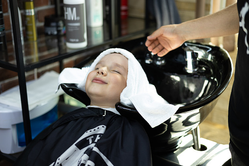 The boy closed his eyes and enjoys while the hairdresser gently wipes his head with a towel in a beauty salon. After washing the hair in the barbershop, the child wipes his head with a fluffy towel..