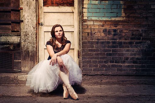 Pretty Ballerina Sitting In Lonely Ally On A Door Stoop smiling and looking away.  Arms crossed at the wrist, legs at the ankle.  Long netted skirt, and two well worn ballet shoes.    Old brick wall background with a faded old door.  Copy Space.