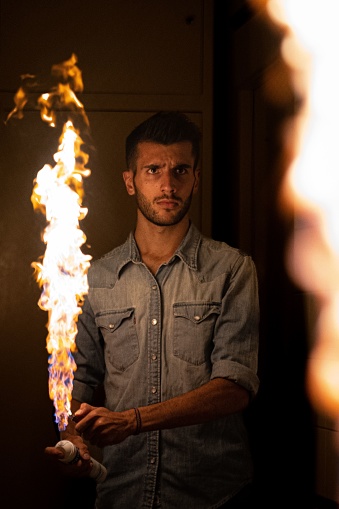An adult male standing in a dimly lit room, holding a burning flame in his hand