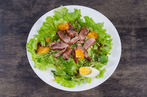 Salad made with boiled duck meat, egg, orange slices and different greens on a white dish on a black surface, top view
