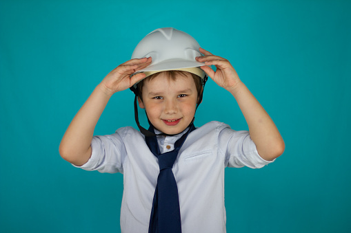 The child adjusts the construction helmet on his head and smiles cutely at the camera isolated on a blue background. A child plays builder smiling and holding on to a construction helmet..