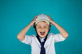 Funny boy child screaming while holding on to a white construction helmet on his head. On a blue background, a boy child in a construction helmet screams holding his head.