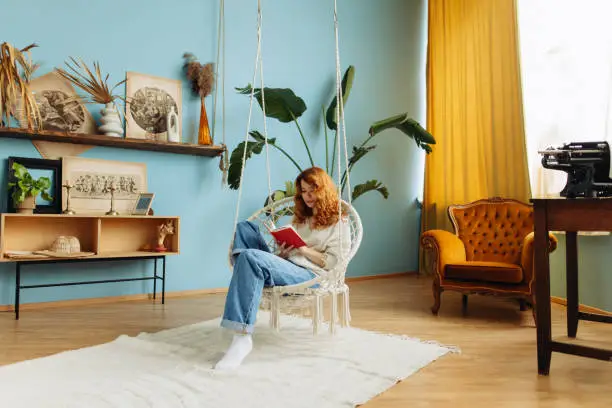 A beautiful young woman with red hair is depicted sitting on a hammock rope swing chair in a bright blue and green colored room. She appears relaxed and engrossed in the pages of a book, creating a serene and peaceful ambiance.