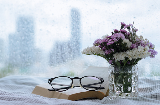 Selective focus on spectacles and book put on bed in morning with rain drop on window. Stay home and relaxing concept.