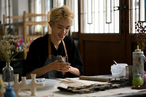 A happy and attractive young Asian gay man enjoys making earthenware, kneading raw clay to sculpt an ornament in a handicraft workshop.