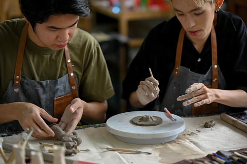 A happy young Asian gay man and his male friend enjoy making clay ornaments in a handicraft workshop together.