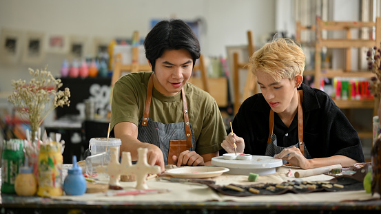 An attractive young Asian gay man enjoys painting a ceramic plate in a creative handicraft workshop with his boyfriend. LGBTQ+, Handicraft workshop concept
