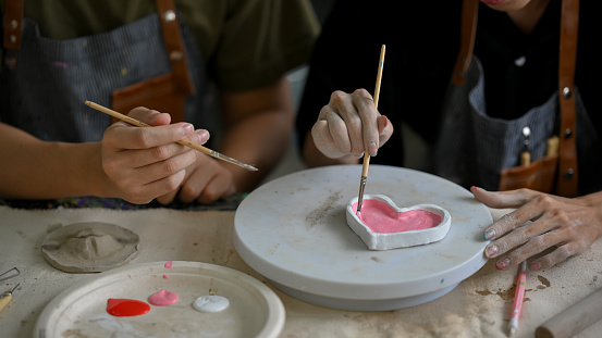 Close-up image of a man enjoys painting a ceramic heart plate in a creative handicraft workshop with his friend. Handicraft workshop concept