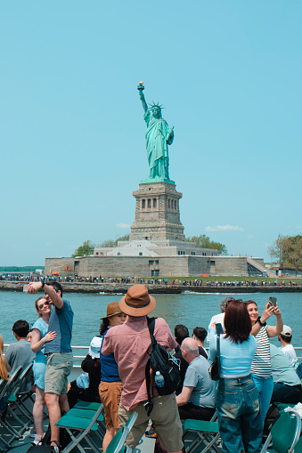New York City, United States - May 22, 2023: Tourists on board of a tour boat browsing in front of the Statue of Liberty, in Liberty Island, New York, United States, on a sunny spring day