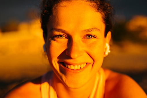 Portrait of young smiling woman sitting on the beach and listening music on headphones before a morning running session at the beach . Female runner at the beach.