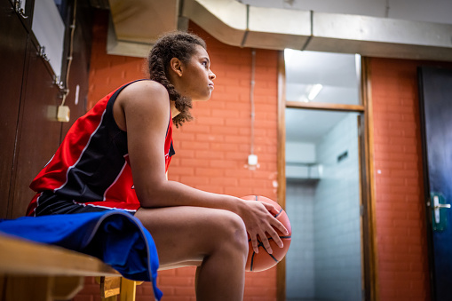Female basketball player holding ball while sitting on bench in sports court during practice.