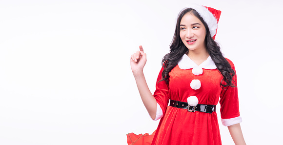 Cheerful surprise asian woman wear Santa Claus dress with Christmas hat use finger pointing present copy space standing on white background. Excited young girl Santa Claus dress merry Christmas winter