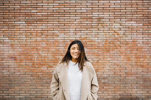 Portrait of a young smiling latin american woman wearing a coat against a brick wall on the street
