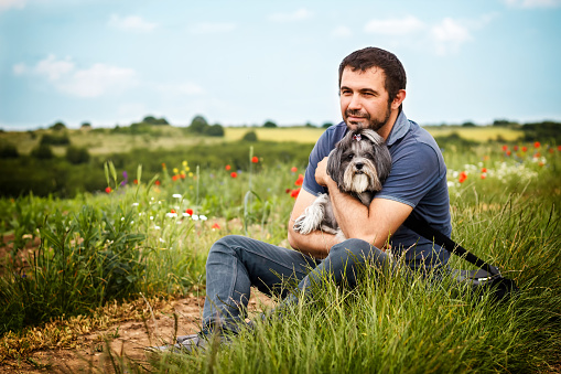 Tranquil and cheerful scene with a white Caucasian bearded man sitting outside in the nature, looking dreamy and hugging his best friend dog. International Dog day. Copy space