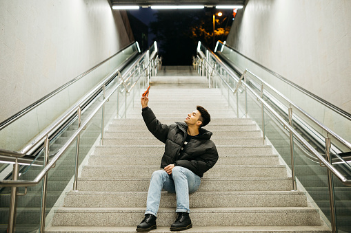 Young man, sitting on stairs at the subway entrance, taking a selfie with a red smartphone