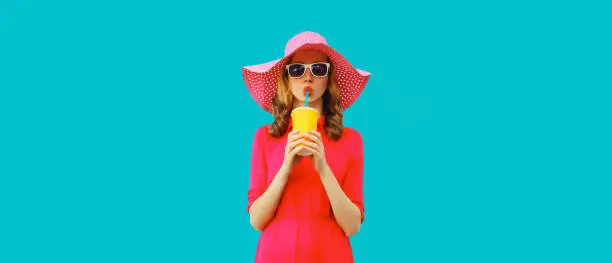 Summer portrait of beautiful young woman drinking fresh juice wearing straw round hat, pink dress on blue background