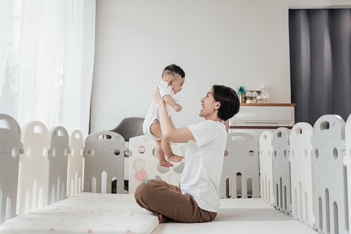 Happiness Asian mother playing with adorable her son in playpen at home. Asian mom lifting and playing newborn baby. Happy asian family healthcare love together mothers day concept.