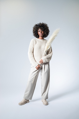 Full length portrait of young woman with afro hairstyle wearing white sweater and pants holding dry pampas grass and looking at camera.