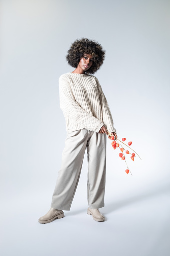 Full length portrait of young woman with afro hairstyle wearing white sweater and pants holding twig of orange physalis and looking at camera.