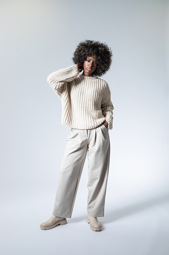 Full length portrait of young woman with afro hairstyle wearing white sweater and pants, looking at camera.