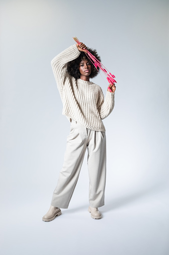 Full length portrait of young woman with afro hairstyle wearing white sweater and pants holding dry red plants and looking at camera.