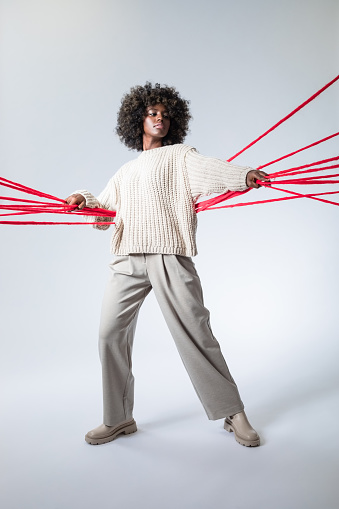 Full length portrait of young woman with afro hairstyle wearing white sweater and pants, standing among red yarn lines, looking at camera.