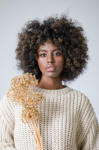 Portrait of young woman with afro hairstyle wearing white sweater holding dried wild garlic flower and looking at camera.