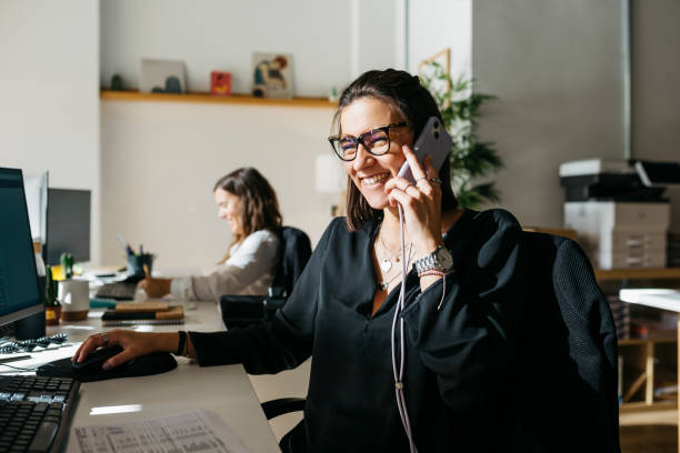 Business woman talking on the phone, communicating with her clients as she works in an office Happy young business woman communicating with her clients over the phone, as she works from an office. small office interior stock pictures, royalty-free photos & images