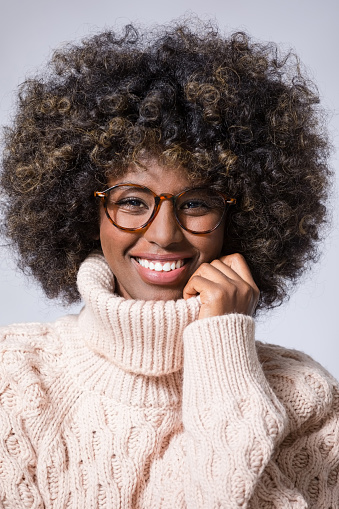 Headshot of young woman with afro hairstyle wearing white wool sweater and eyeglasses,  smiling at camera.