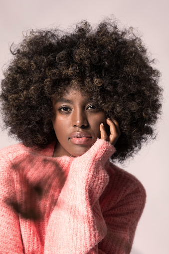 Headshot of young woman with afro hairstyle wearing pink wool sweater, looking at camera.