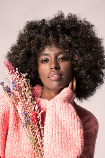 Headshot of young woman with afro hairstyle wearing pink wool sweater, holding dry flowers and looking at camera.