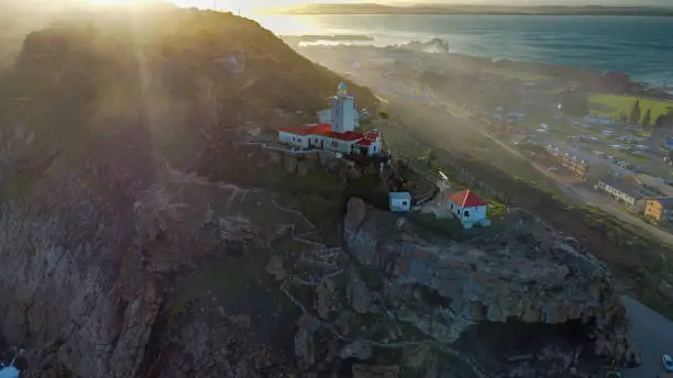 Aerial shot of the historic Cape St. Blaize lighthouse in Mossel bay, constructed in 1864 standing 14 meters tall during sunset with a view of the ocean and the town in the distance.