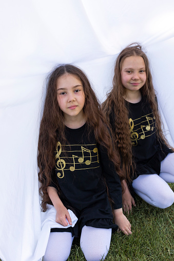 Two Little Girls, Sisters With Long Hair Having Fun Under Sheet On Grass in Park, Making Tent. Caucasian Asian Siblings Enjoy Time Together. Family Love And Care, True Friendship. Vertical Plane