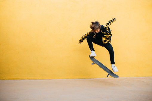 Portrait of Teenager boy jumping with an skateboard in front of a yellow wall, on his way to school