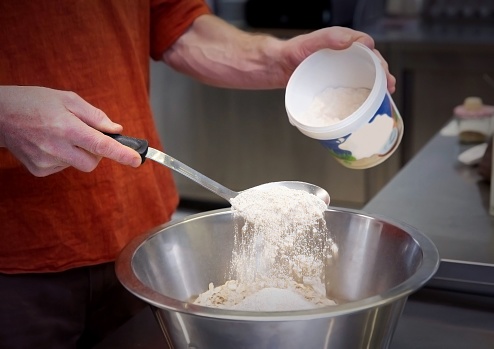 A close-up of a person's hands adding a scoop of white flour to a bowl of dough.